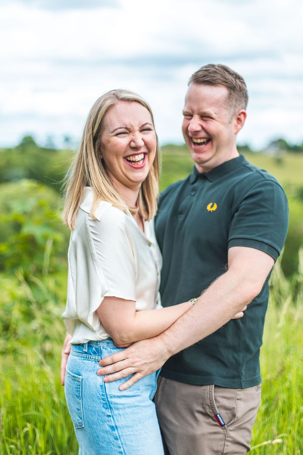 A happy couple laughs together while embracing in a sunny green field on a summer day.