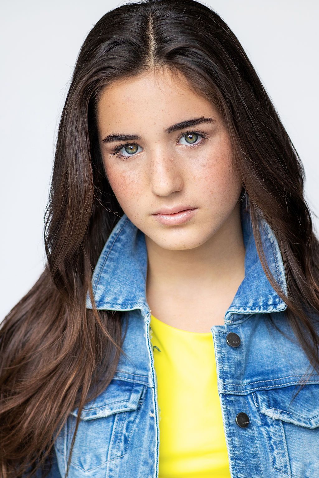 Portrait shot against white background wearing denim jacket and yellow top with long dark hair.