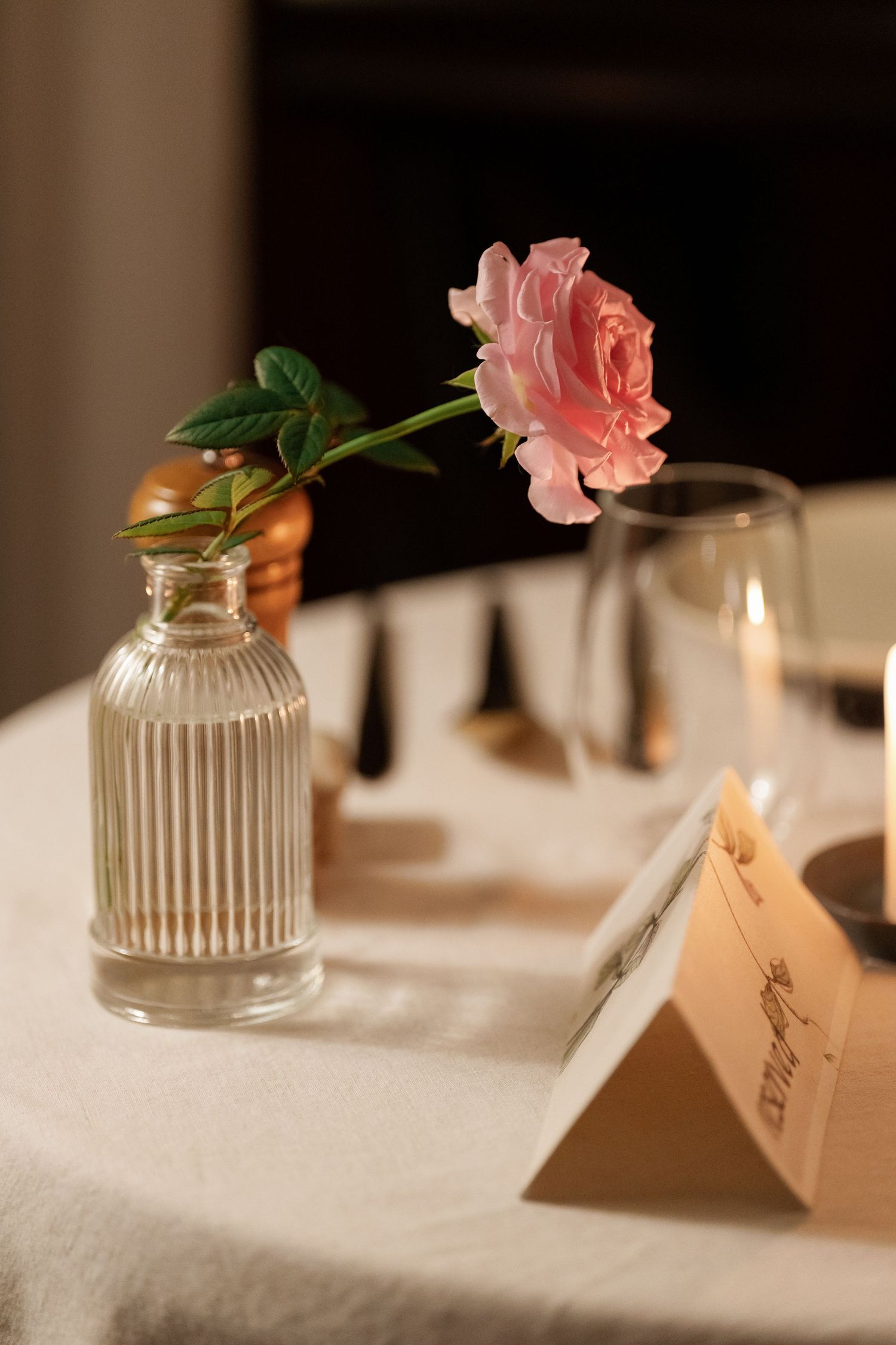 A pink rose in a glass vase sits on a white tablecloth with a candlelit glow and place card in the background.