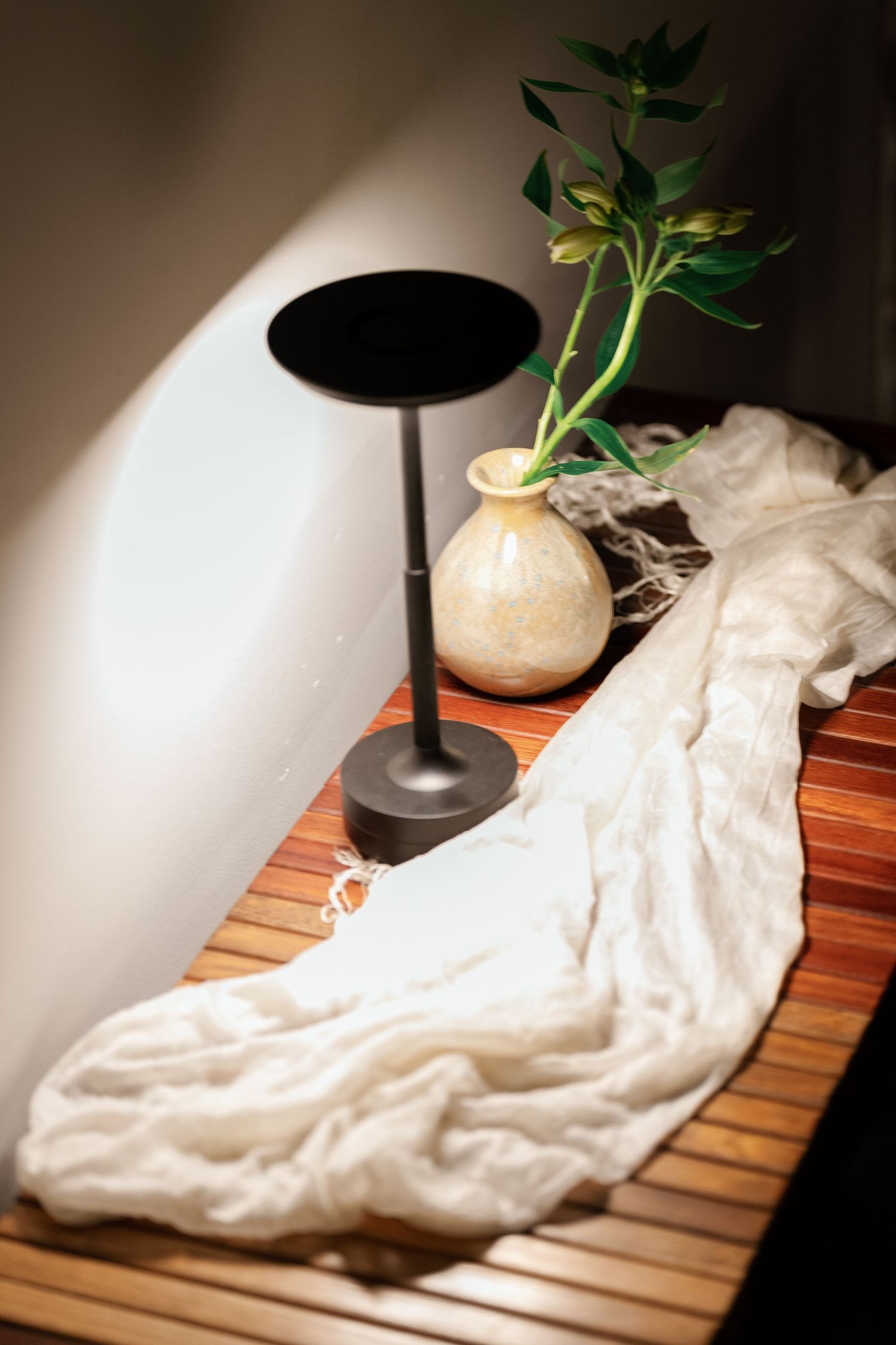 A black modern table lamp and beige vase with green plant sit on wooden slats beside flowing white fabric.