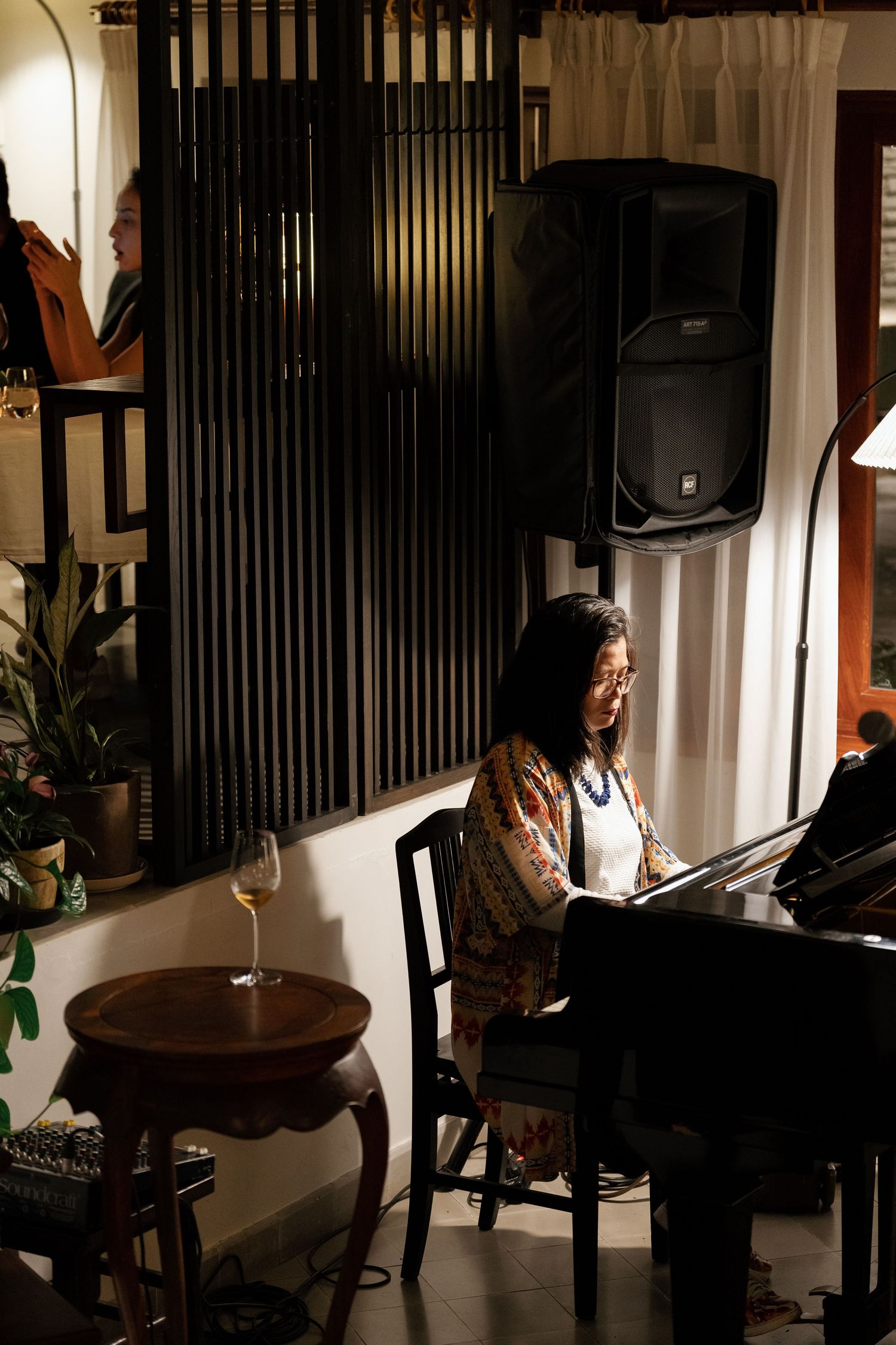 A musician performs at a piano in a dimly lit lounge setting with speakers mounted on the wall and wine glasses on tables.