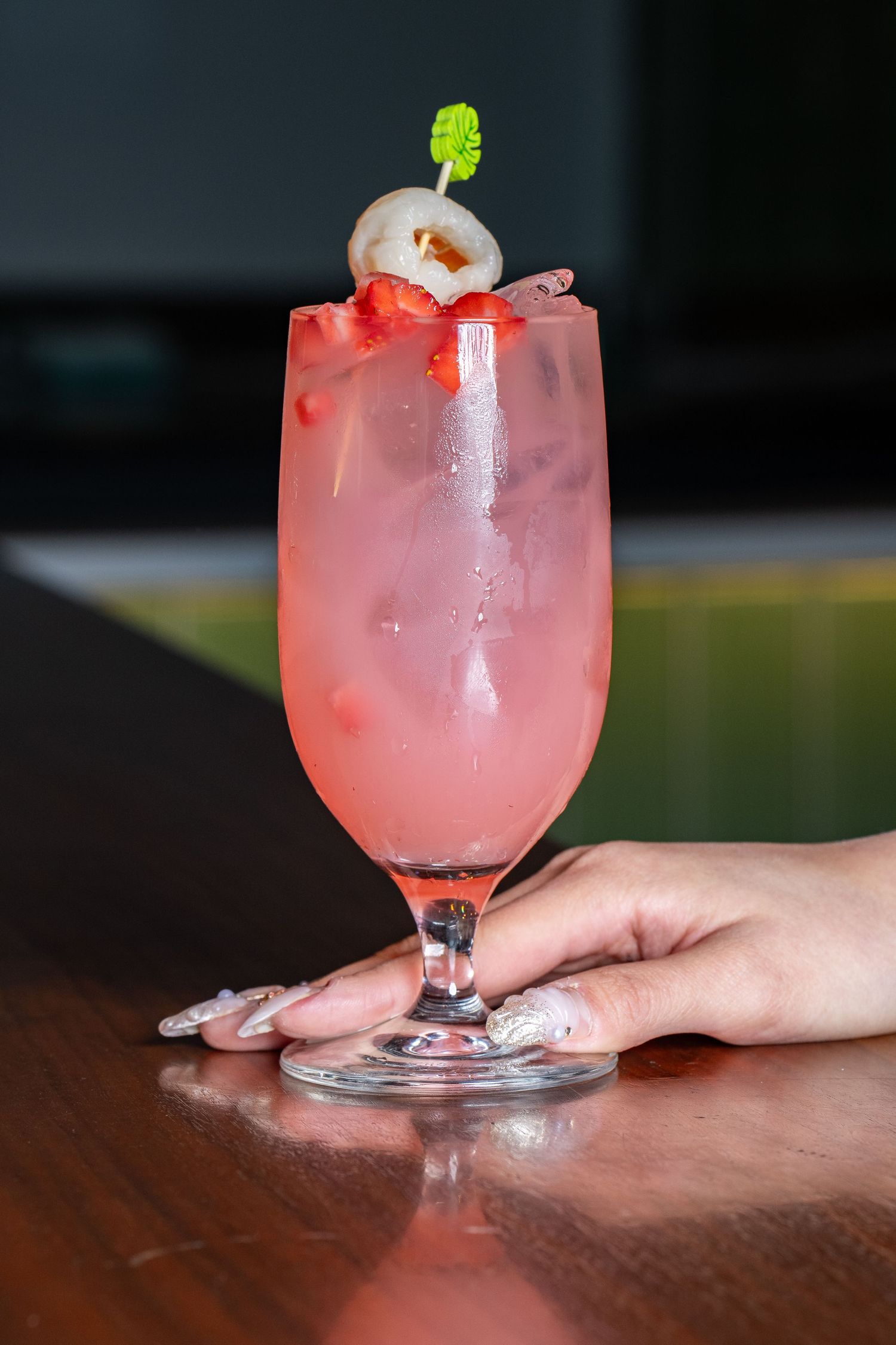 Pink cocktail in a stemmed glass garnished with a lychee and herb sprig, served on a wooden bar top.