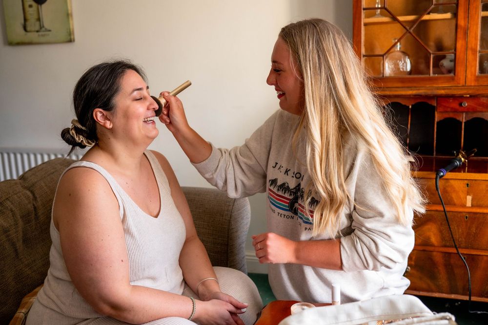 Professional makeup artist applying cosmetics to a client seated on a gray couch in a home setting.