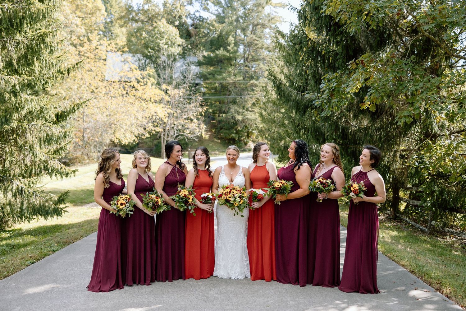 Wedding party in burgundy and red bridesmaid dresses standing together on garden path among evergreen trees.