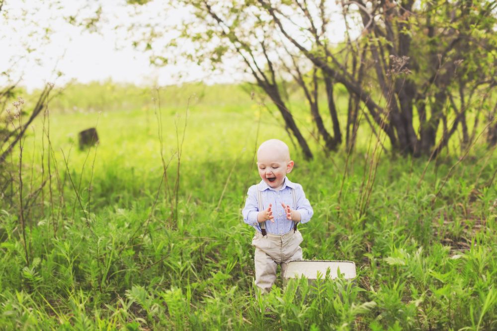 A baby crawls through tall green grass in a sunny meadow, wearing light-colored clothing.