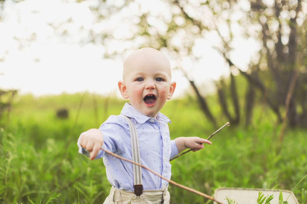 A joyful toddler in suspenders and light blue shirt holds a stick while playing outdoors in a sunny green field.