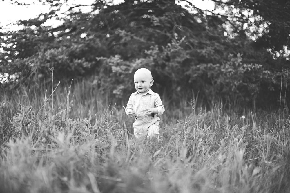 A black and white photograph shows a small child standing among tall grass with trees in the background.