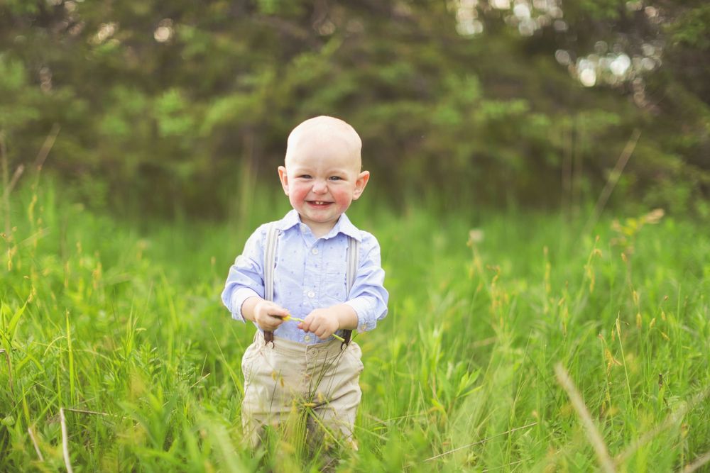 Happy toddler wearing suspenders and blue shirt stands in lush green spring meadow.