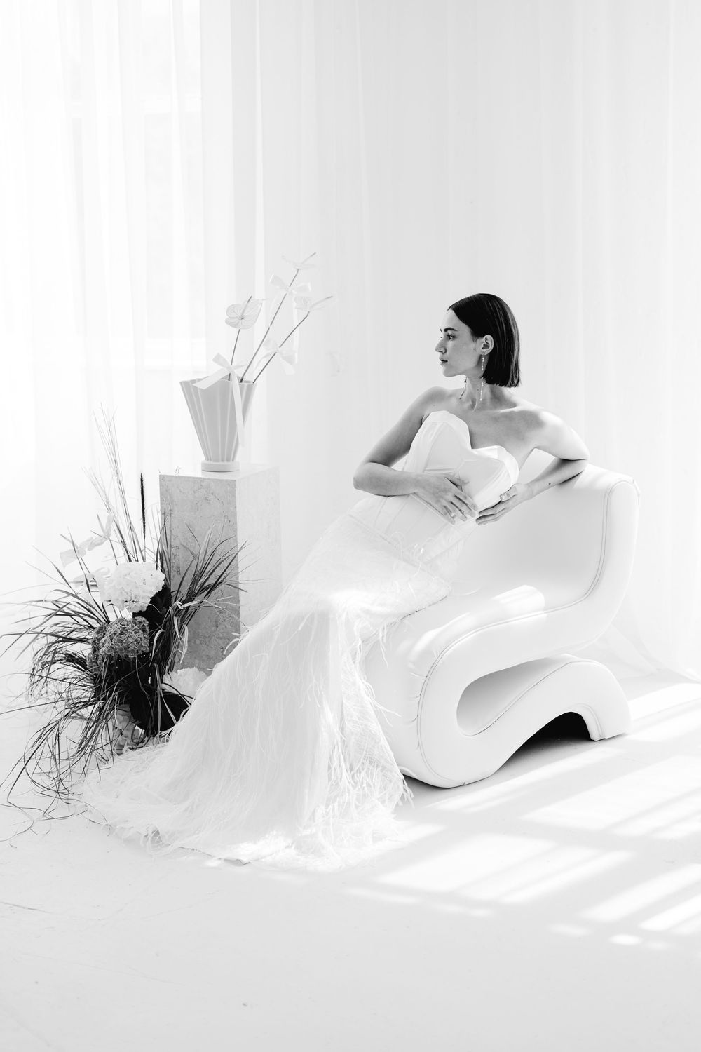 A figure in a white gown poses elegantly on a modern curved chair against a bright white backdrop.