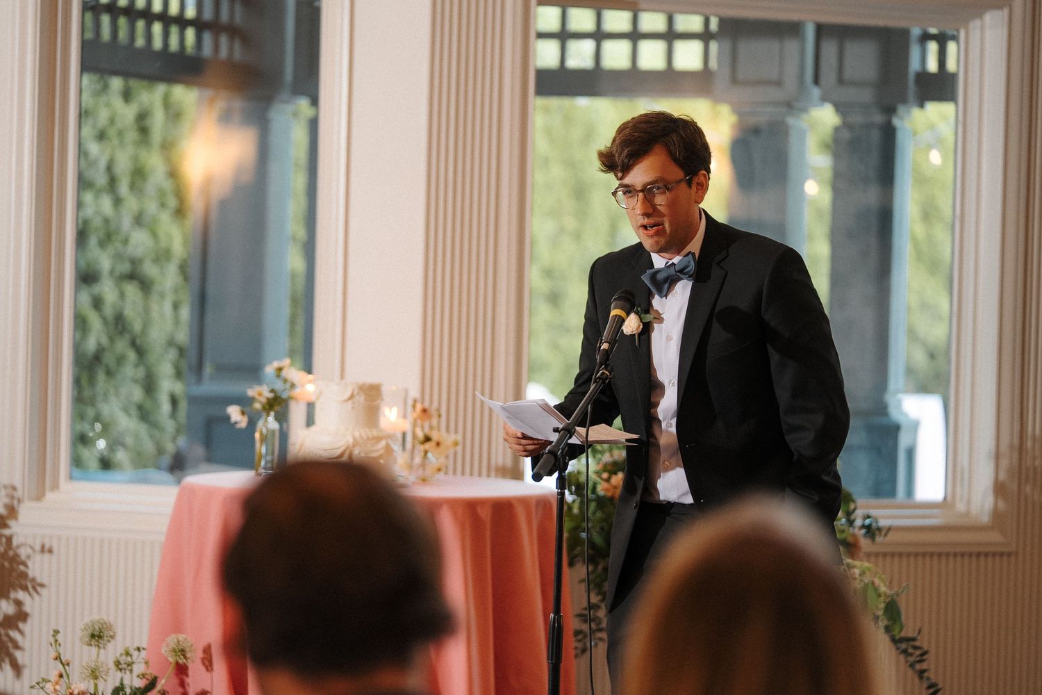 A person in a black tuxedo and bow tie gives a speech at a wedding reception with pink table decor.
