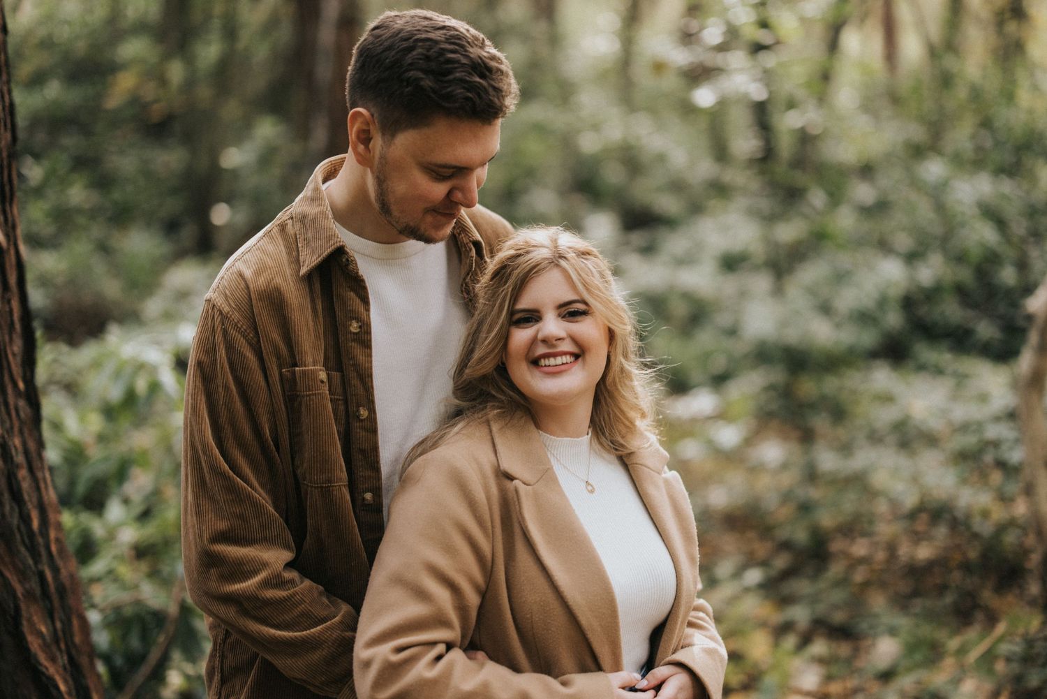 A couple embraces in a romantic autumn forest setting wearing brown coats and smiling warmly. Captured by Lincolnshire photographers.