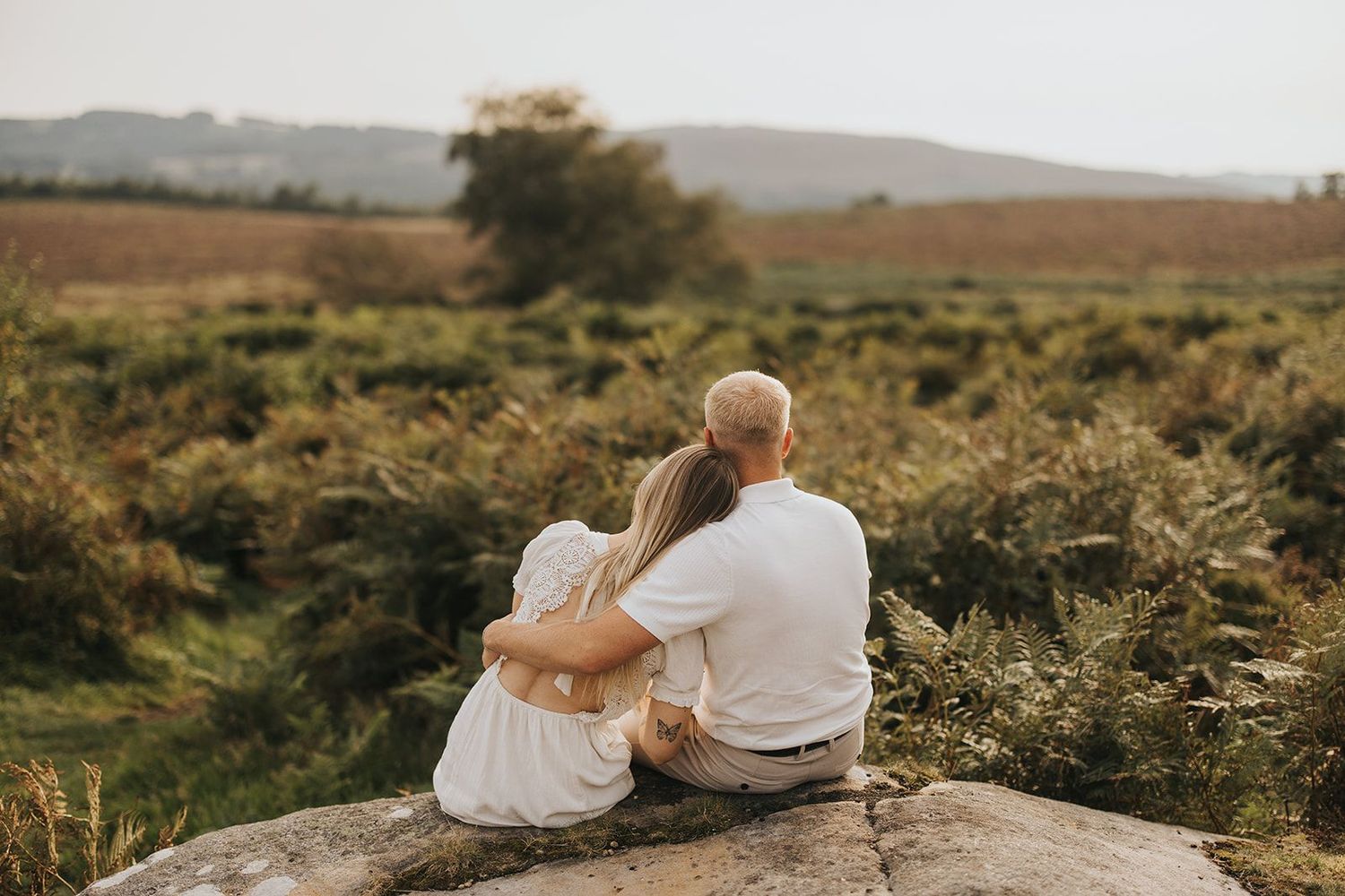 Two people sit together on a rocky outcrop overlooking scenic countryside during golden hour sunset. Captured by Lincolnshire photographers.