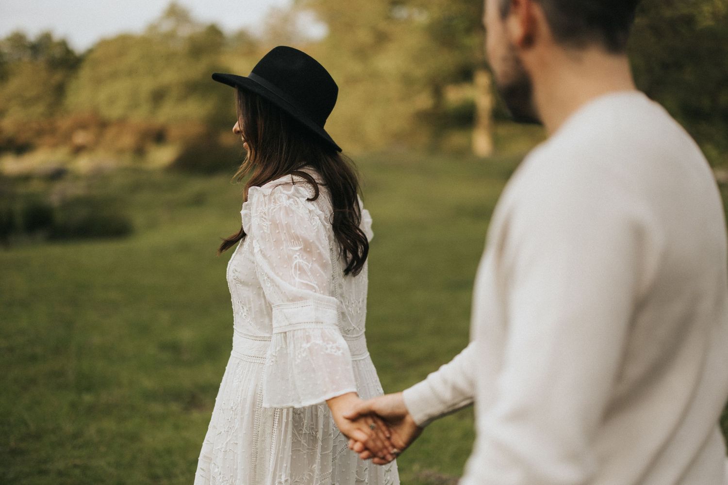 A couple walks hand in hand outdoors, with the lady wearing a white lace dress and black hat. Captured by Lincolnshire photographers.