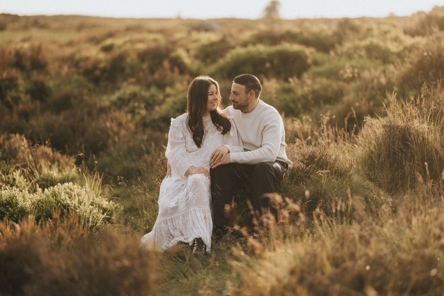 A newly engaged couple sit together on grass during golden hour in a romantic outdoor setting. Captured by Lincolnshire photographers.