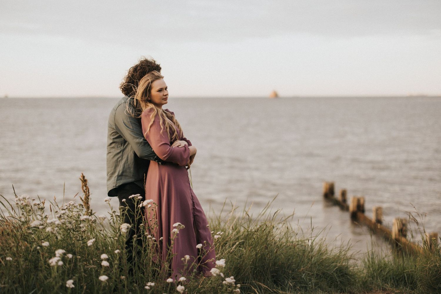 A couple walking along Cleethorpes beach shoreline with wooden posts in the water during golden hour. Captured by Lincolnshire photographers