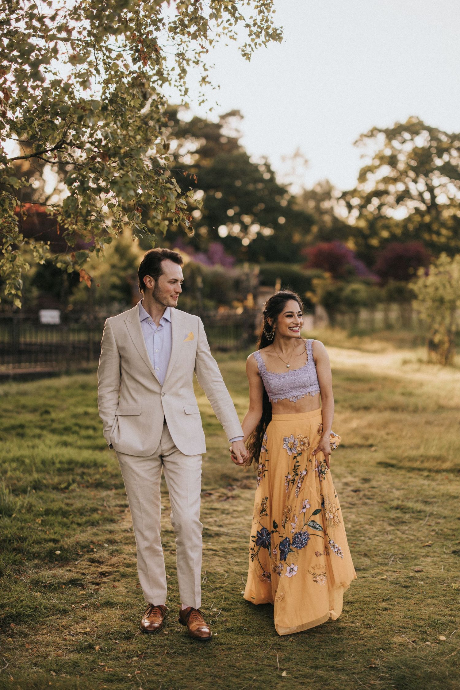 A newly engaged couple walks hand in hand through a sunlit field. Captured by Lincolnshire photographers.