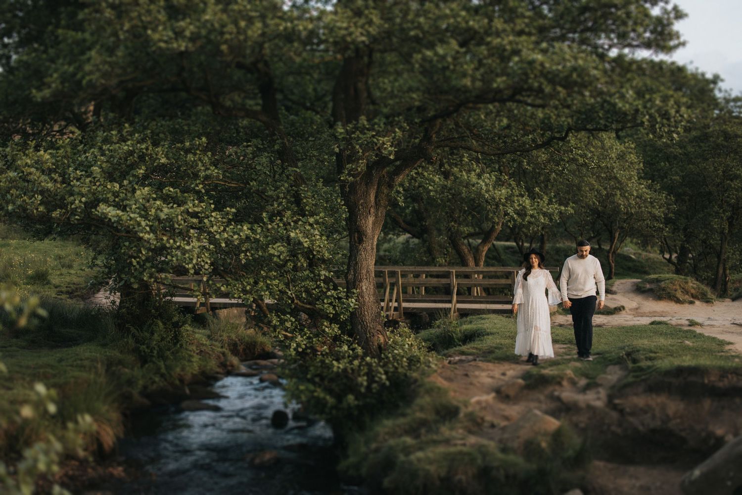 Two people walk along a rustic wooden bridge over a flowing stream surrounded by lush green trees in a scenic outdoor setting.