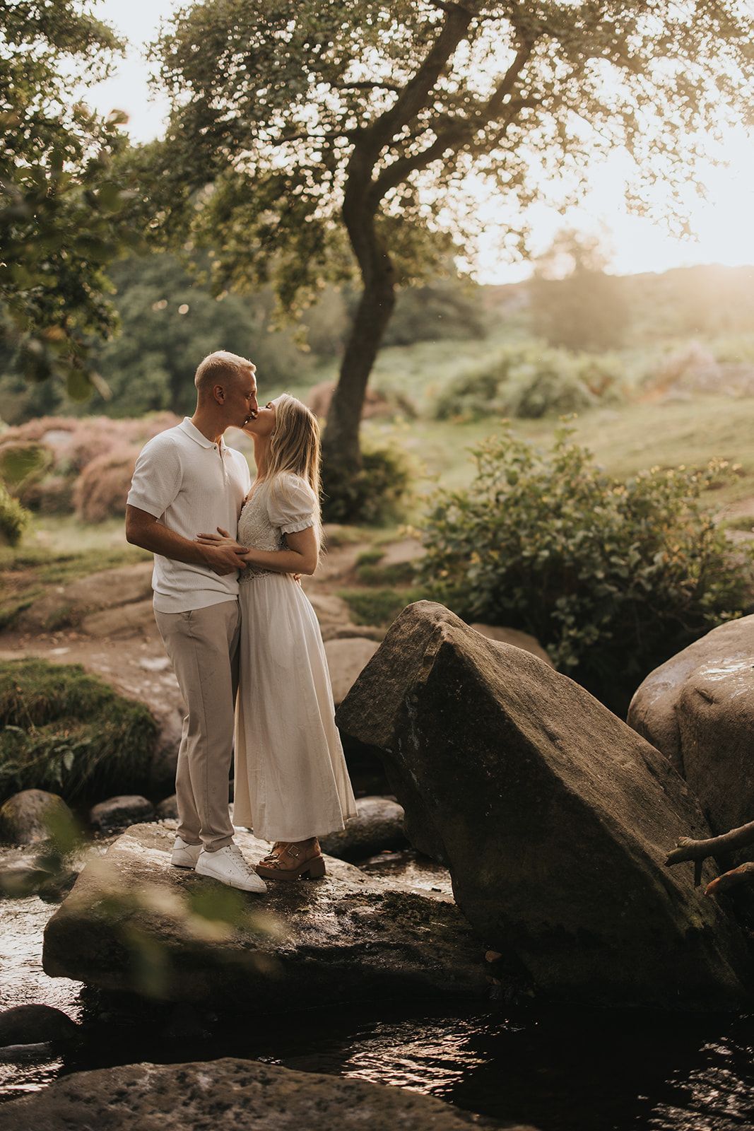 A couple embraces on rocky terrain during golden hour sunset with a tree silhouetted in the warm summer background.