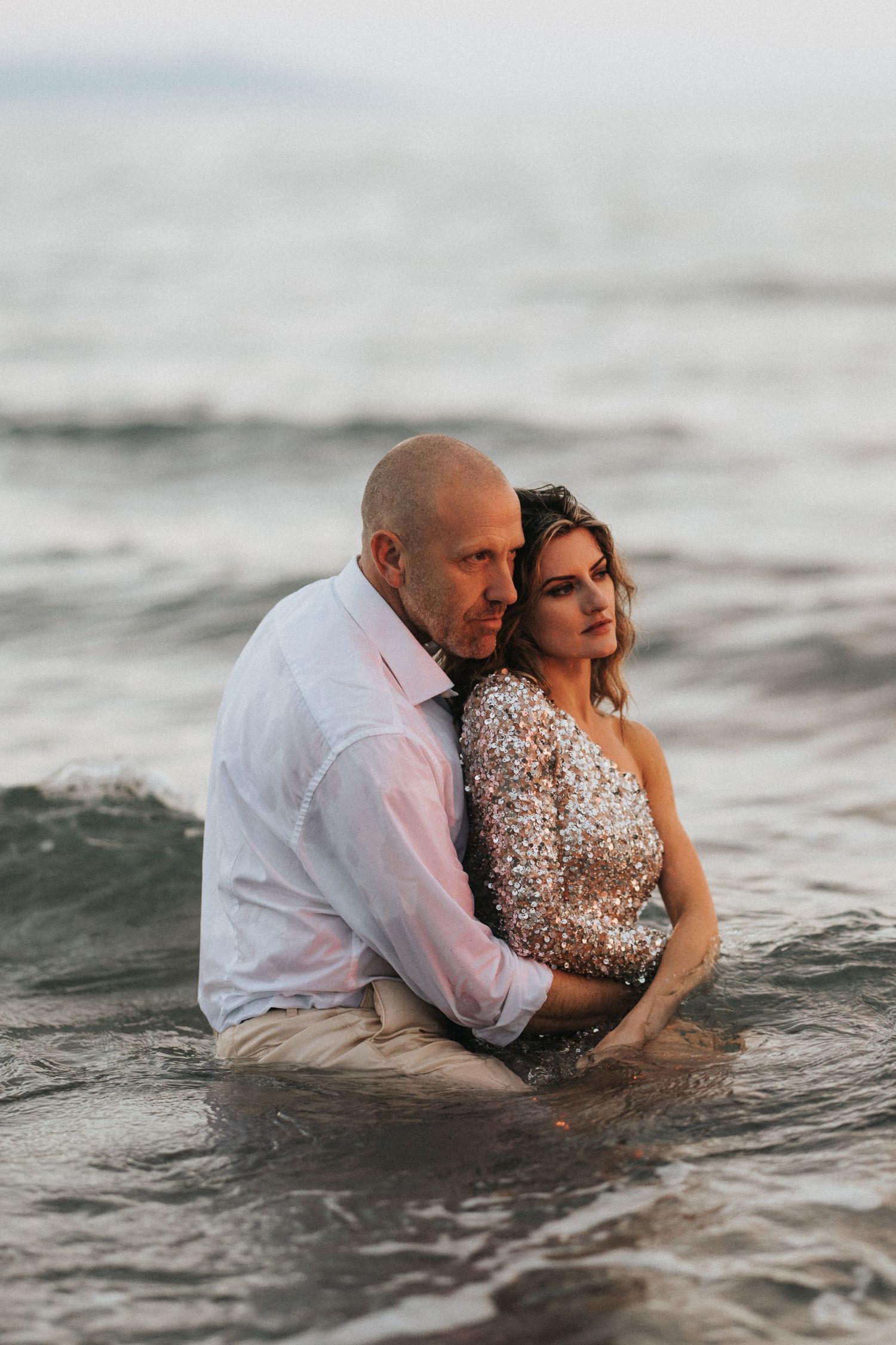 A couple embraces while sitting together in the ocean waves during a sunset photoshoot. Captured by Lincolnshire photographers.
