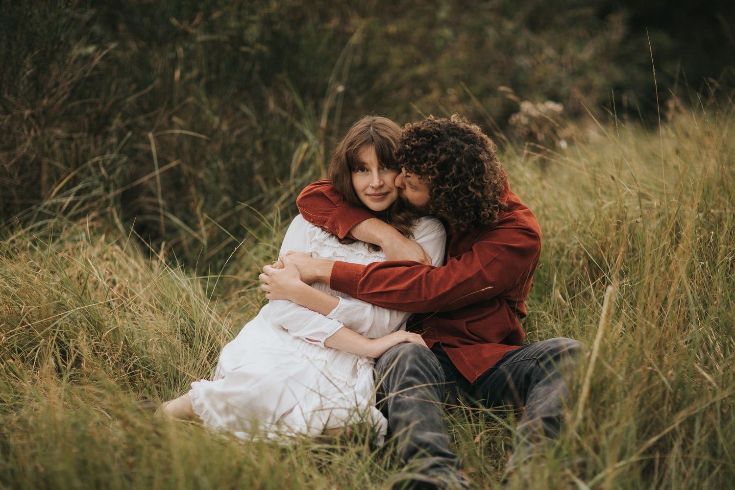 A couple in rustic attire embrace while sitting in a field of tall grass on an autumn day. Captured by Lincolnshire photographers.