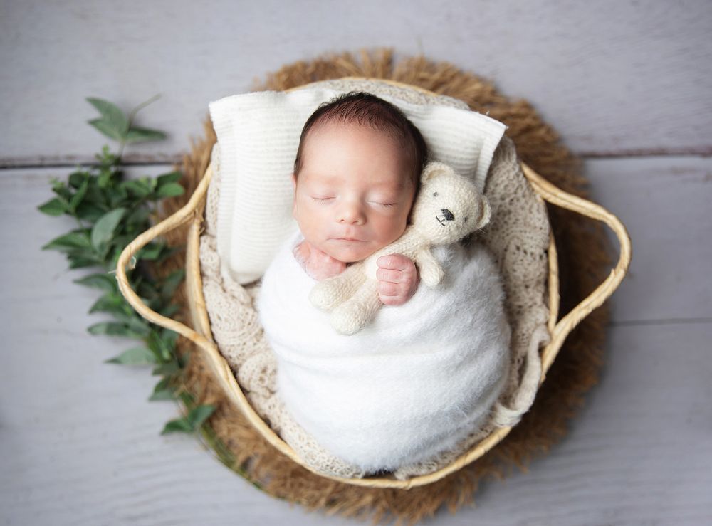 A sleeping newborn swaddled in white blanket rests in a woven basket with plush teddy bear and greenery accents.