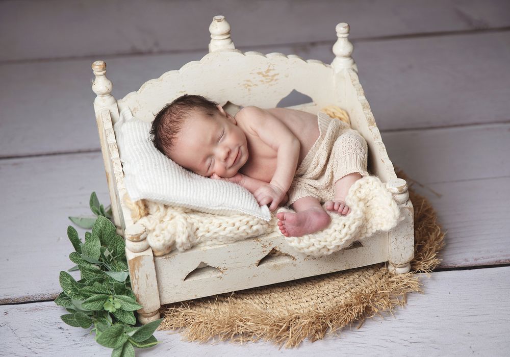 A sleeping newborn rests peacefully in a vintage white wooden bed with lace bedding and greenery decor.