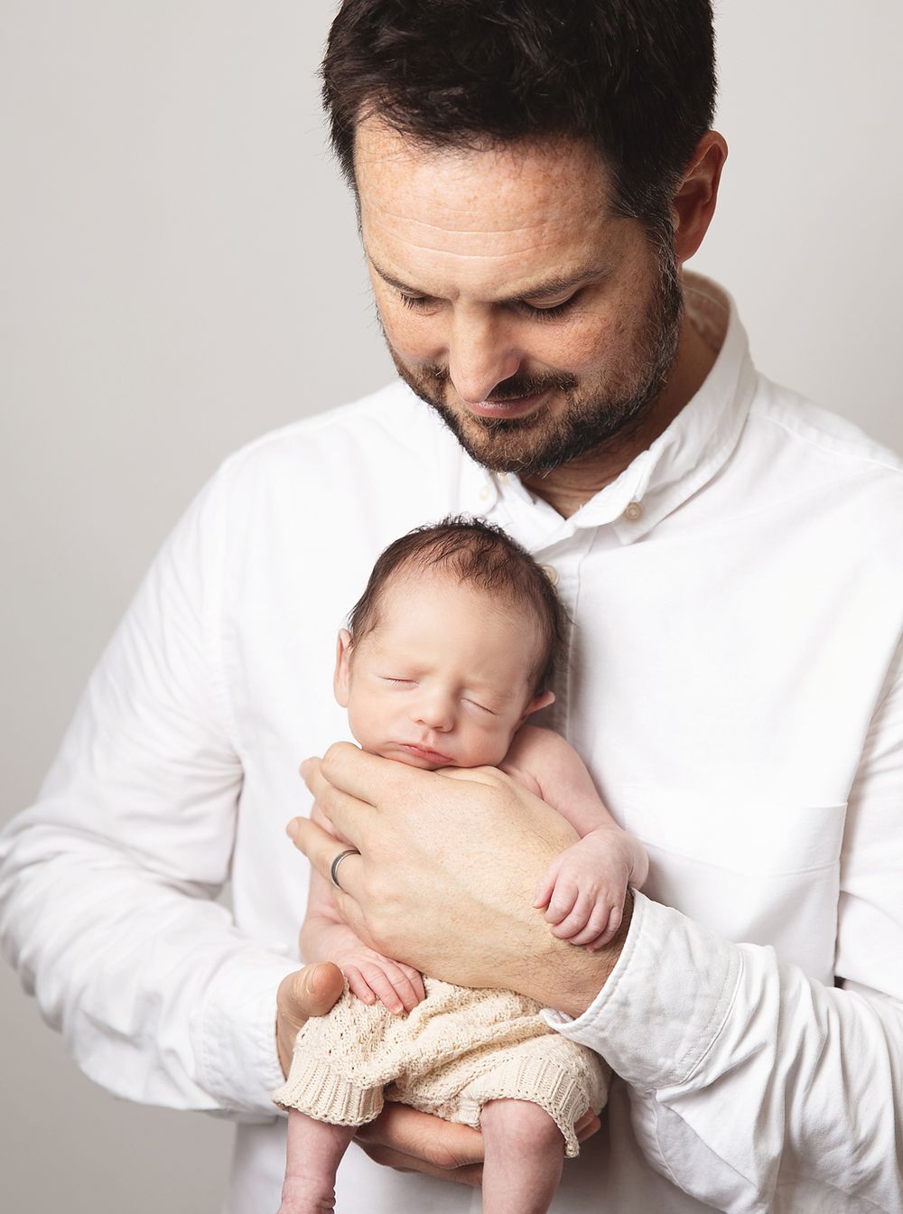Loving parent in white shirt holding sleeping newborn baby in knitted shorts against neutral background.