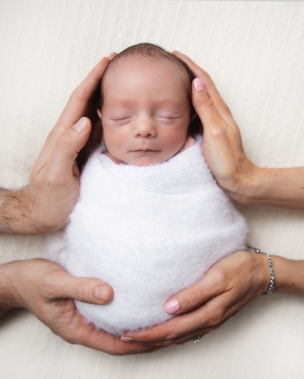 Gentle hands cradle a sleeping newborn swaddled in soft white blanket showing tender care and parental love.
