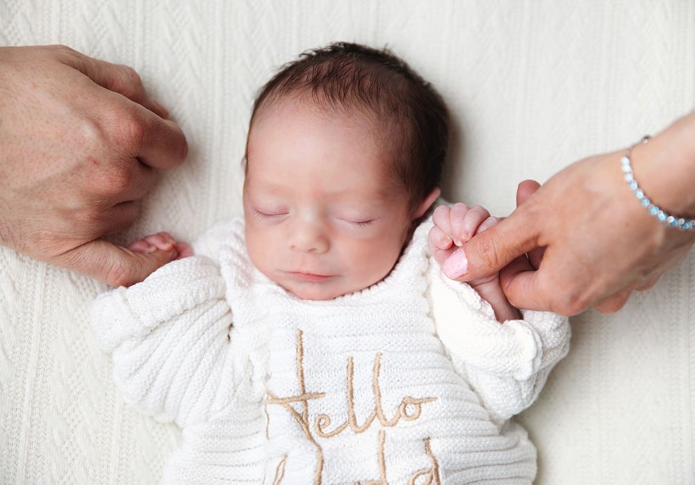 Small baby lies on white bedding in crisp white cotton onesie while being gently touched by reaching hands.