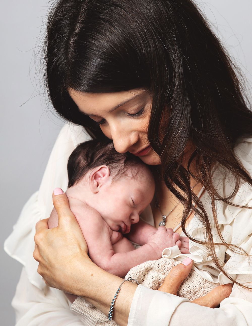 A mother in white holds her sleeping newborn baby tenderly against her chest in an intimate portrait.