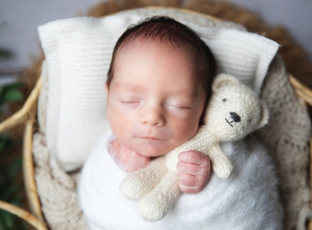 Sleeping newborn in white swaddle nestled in basket with small plush teddy bear toy.