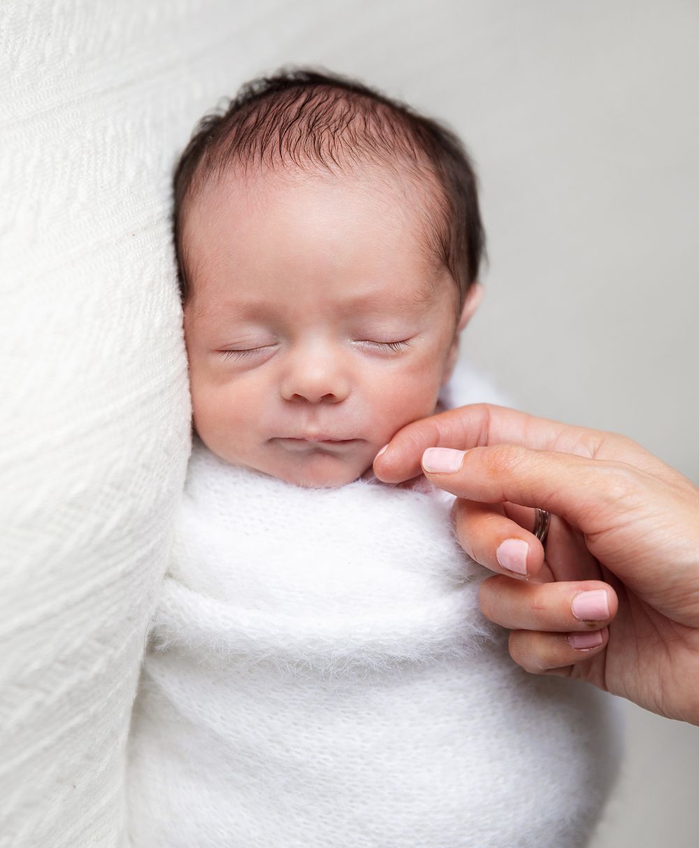 A peaceful sleeping newborn baby swaddled in white fabric with a gentle hand touching their cheek.