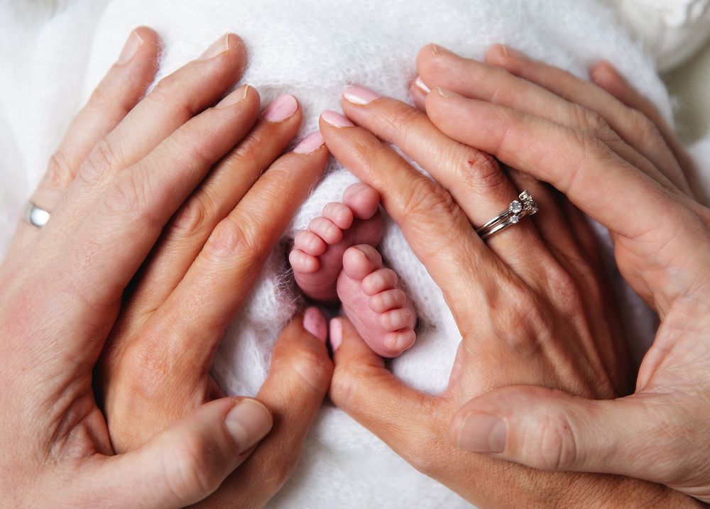 Parent hands gently cradle tiny newborn baby feet wrapped in a soft blanket, with an engagement ring visible.