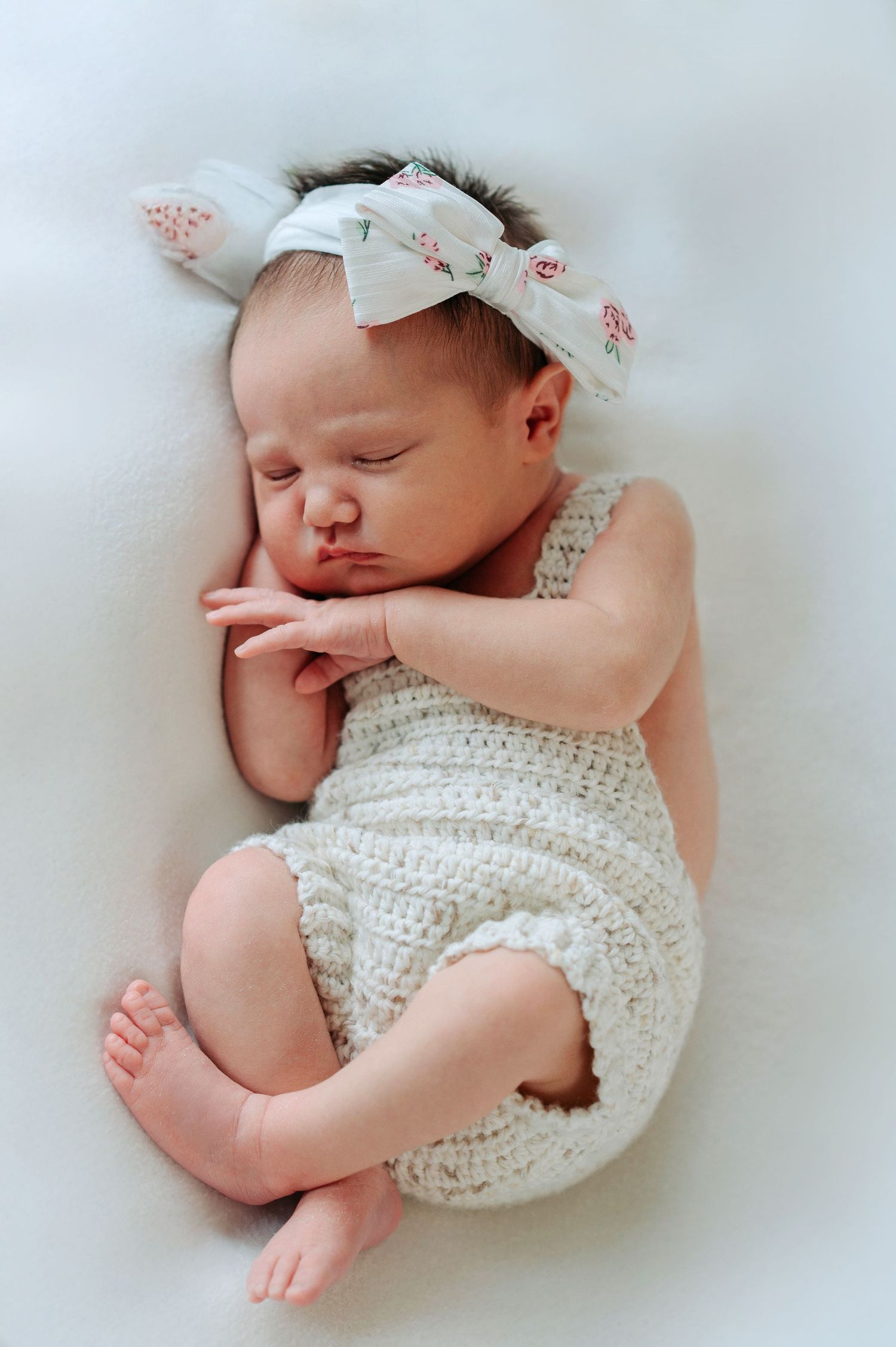 Sleeping newborn wearing white knit romper and floral headband resting peacefully on white background.