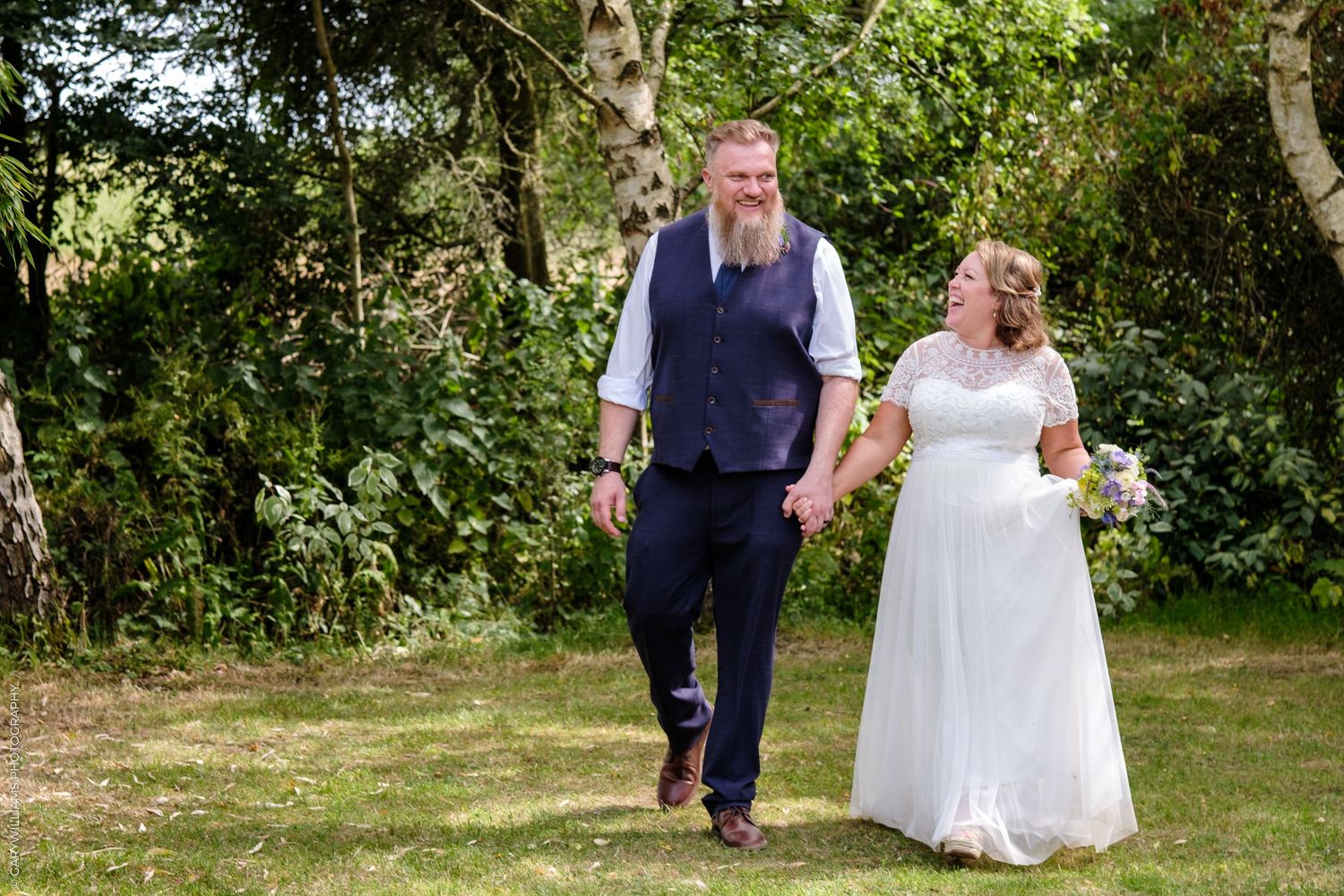 A couple holds hands while walking together on a garden path surrounded by lush greenery and trees.