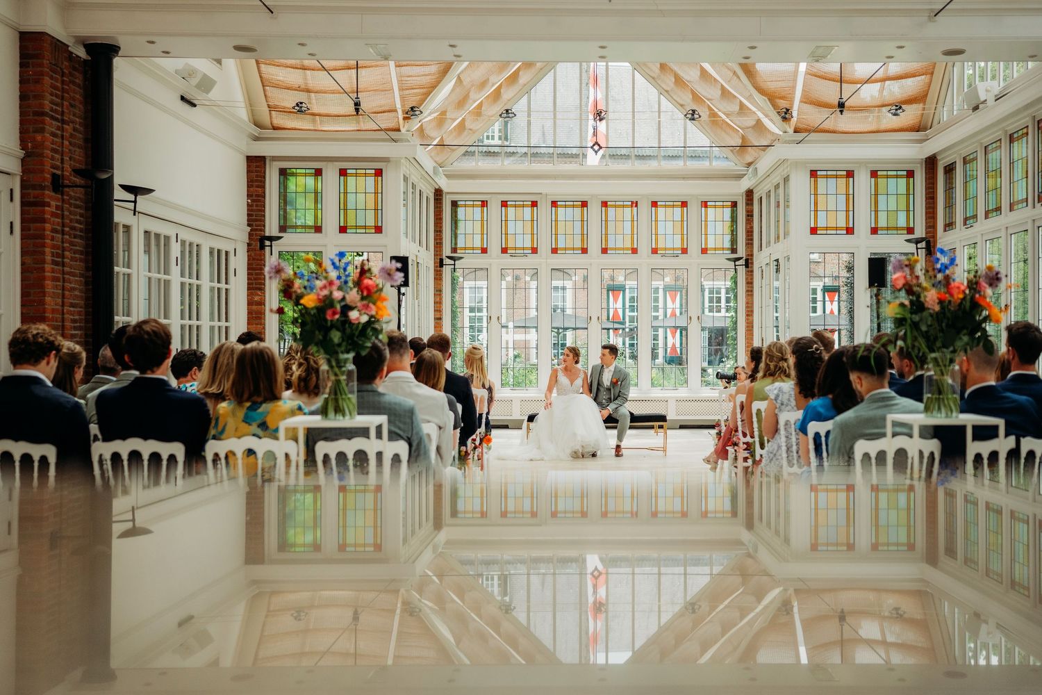 Elegant wedding venue with ornate white decor features long reception tables and dramatic floor-to-ceiling windows.