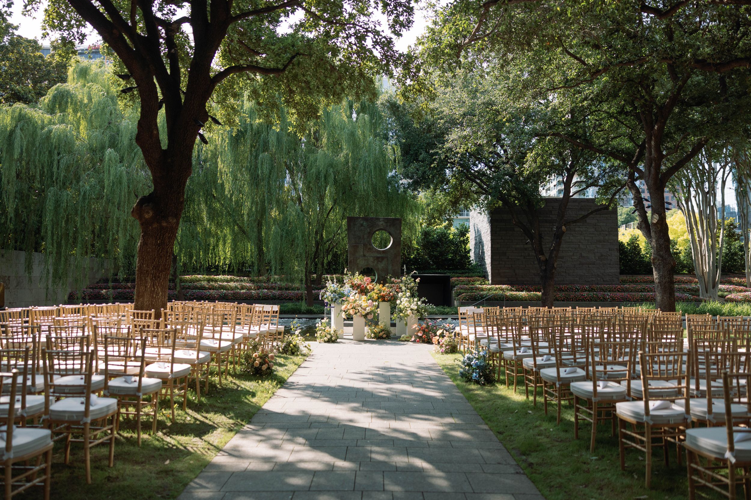Outdoor garden wedding ceremony setup with wooden chairs and willow trees creating a romantic natural backdrop.