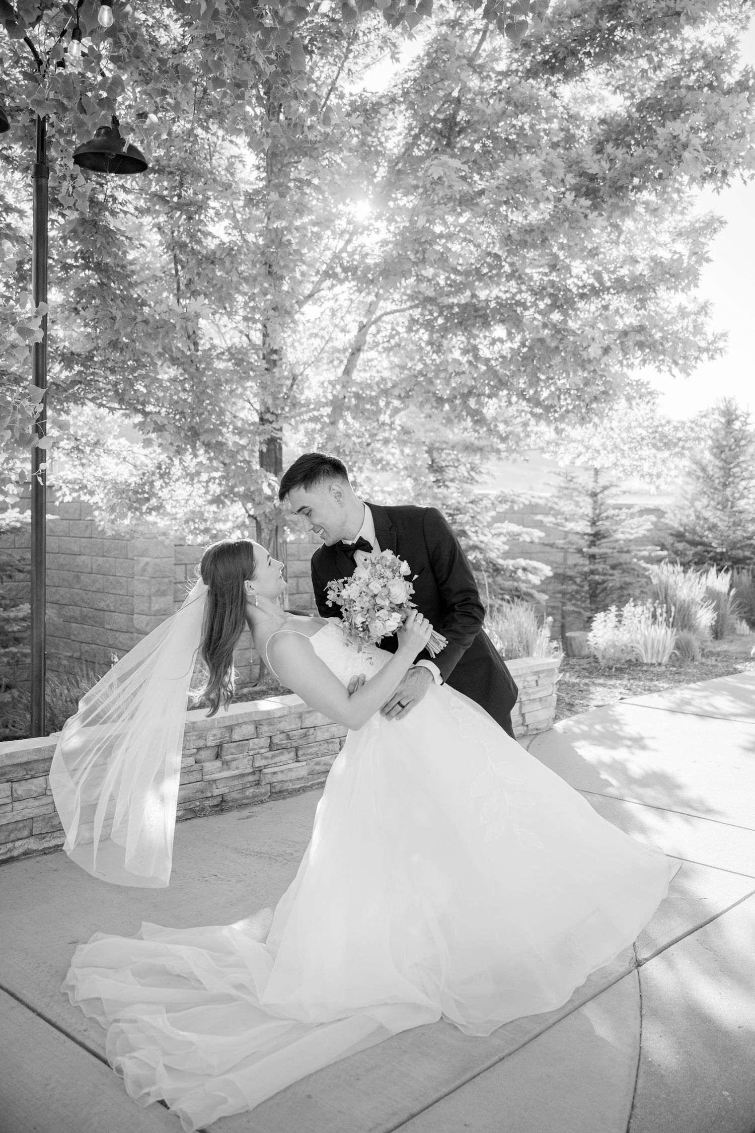 A romantic black and white wedding moment captured at an outdoor venue with trees and stone details in the background.