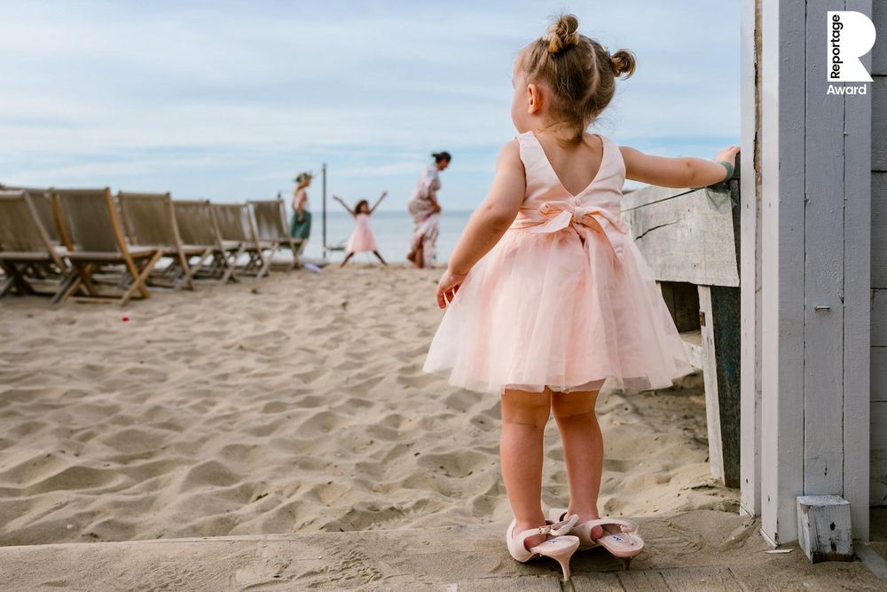 Small child in a pink tulle dress playing in the sand near rustic beach cabanas.