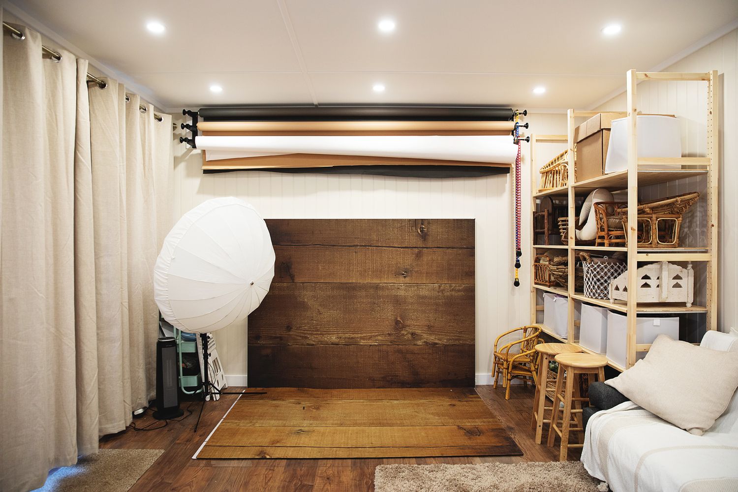 Professional photography studio with wooden backdrop, lighting equipment, white curtains and storage shelves.