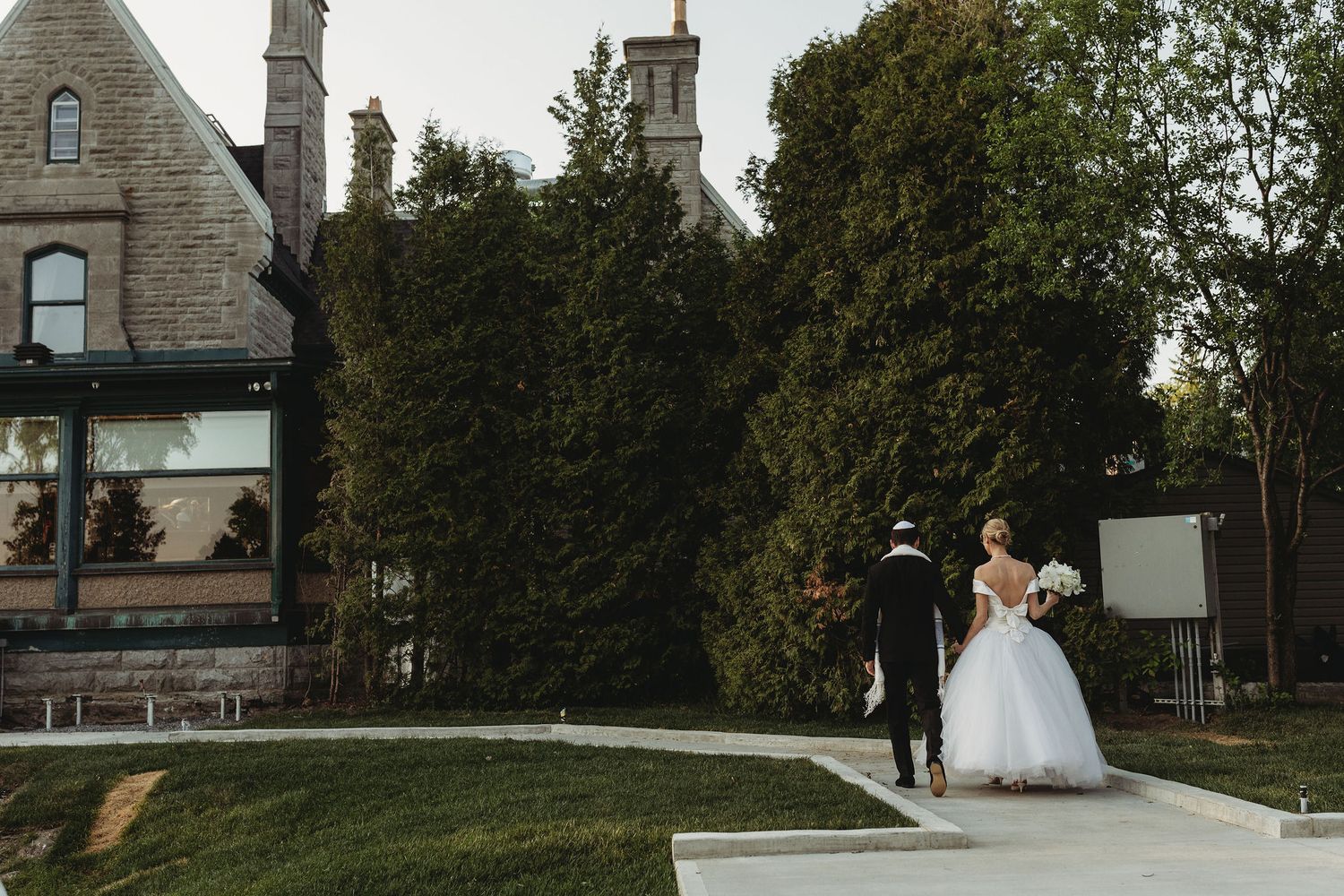 A couple walks together on a stone path outside a historic stone building on their wedding day.
