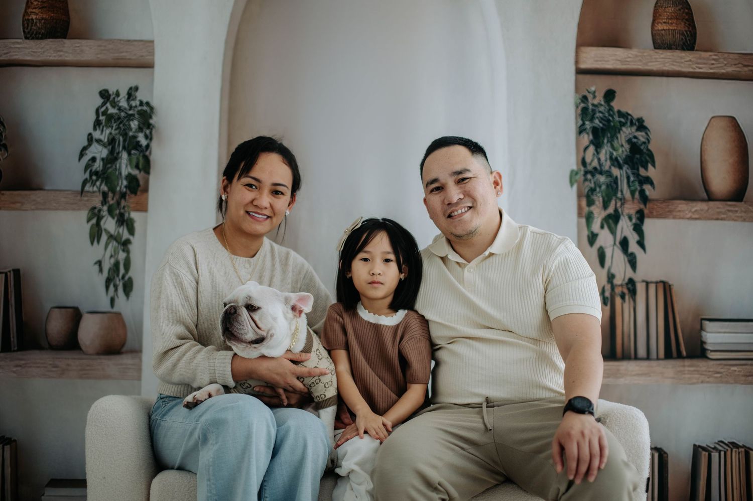 A family sits together on a beige couch with built-in shelving and potted plants in the background.