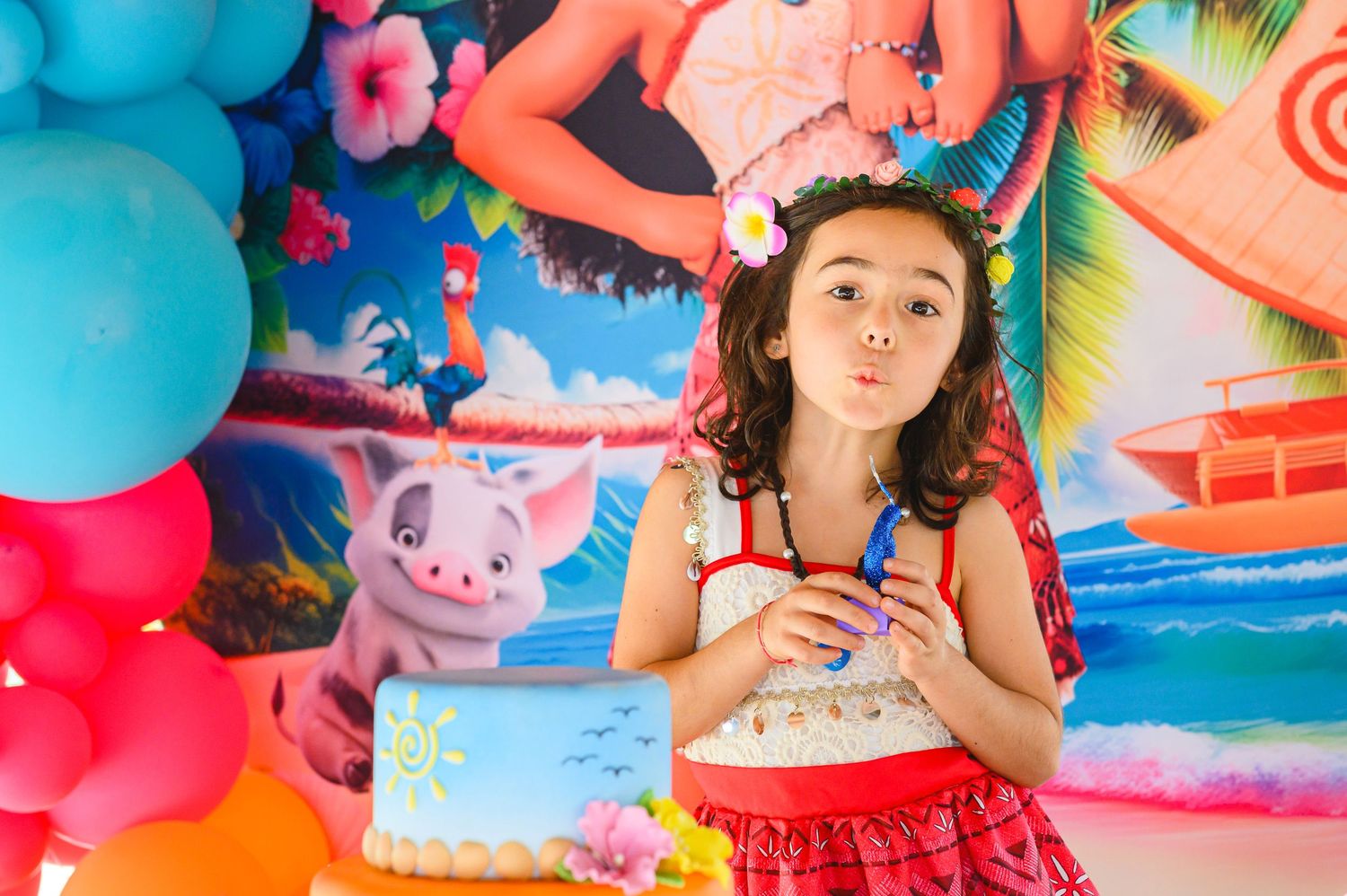 Colorful birthday party scene with balloons, tropical backdrop, and someone in a red skirt holding a drink.