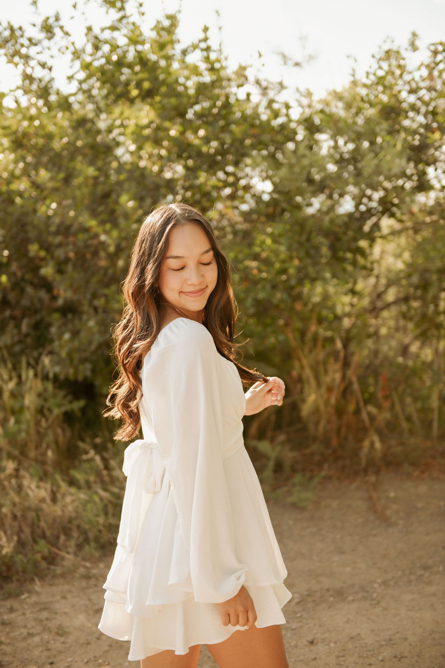 A person in a flowing white dress poses outdoors on a sunlit dirt path with greenery in the background.