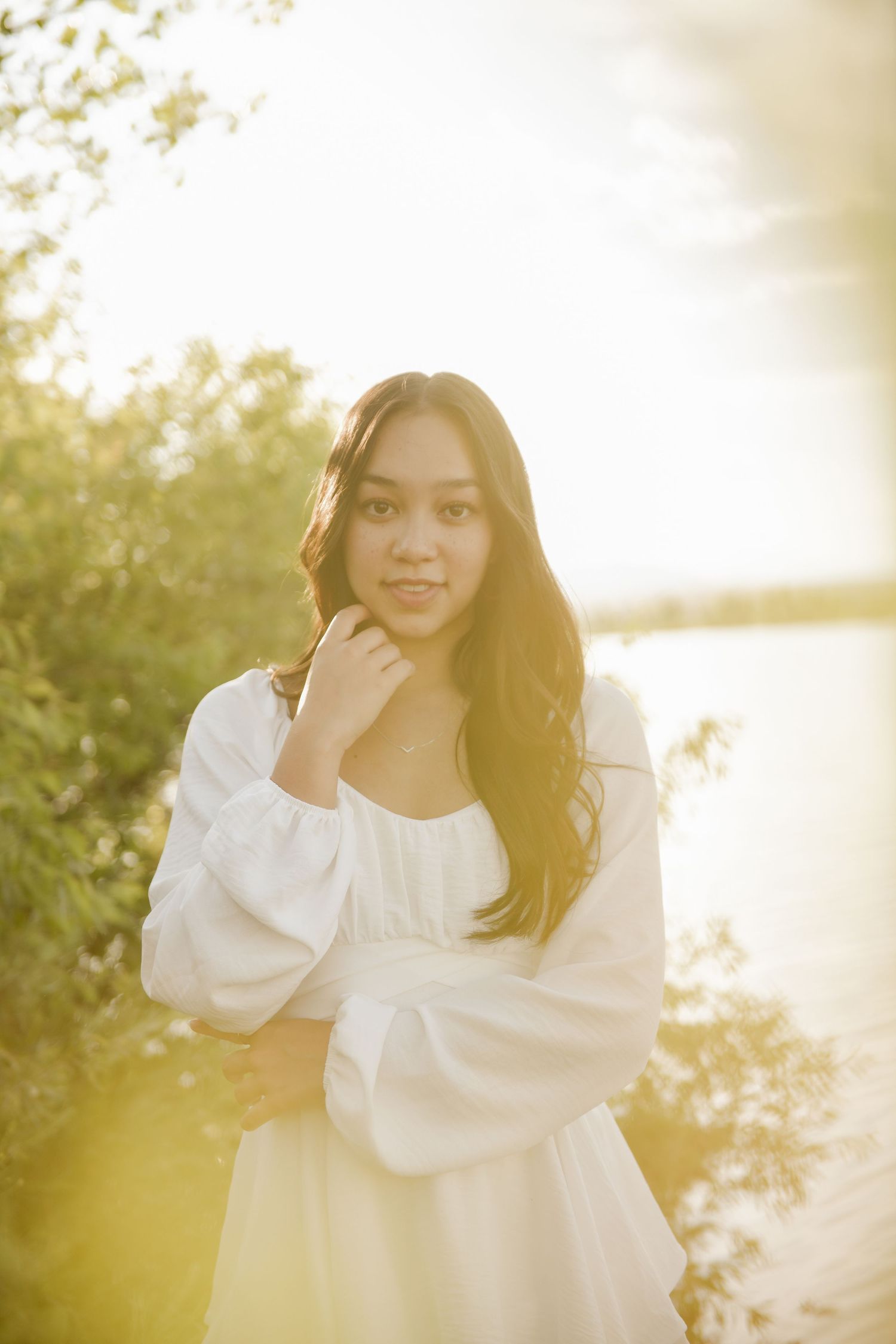 A dreamy portrait in a white dress against golden sunlight filtering through trees by a waterfront.