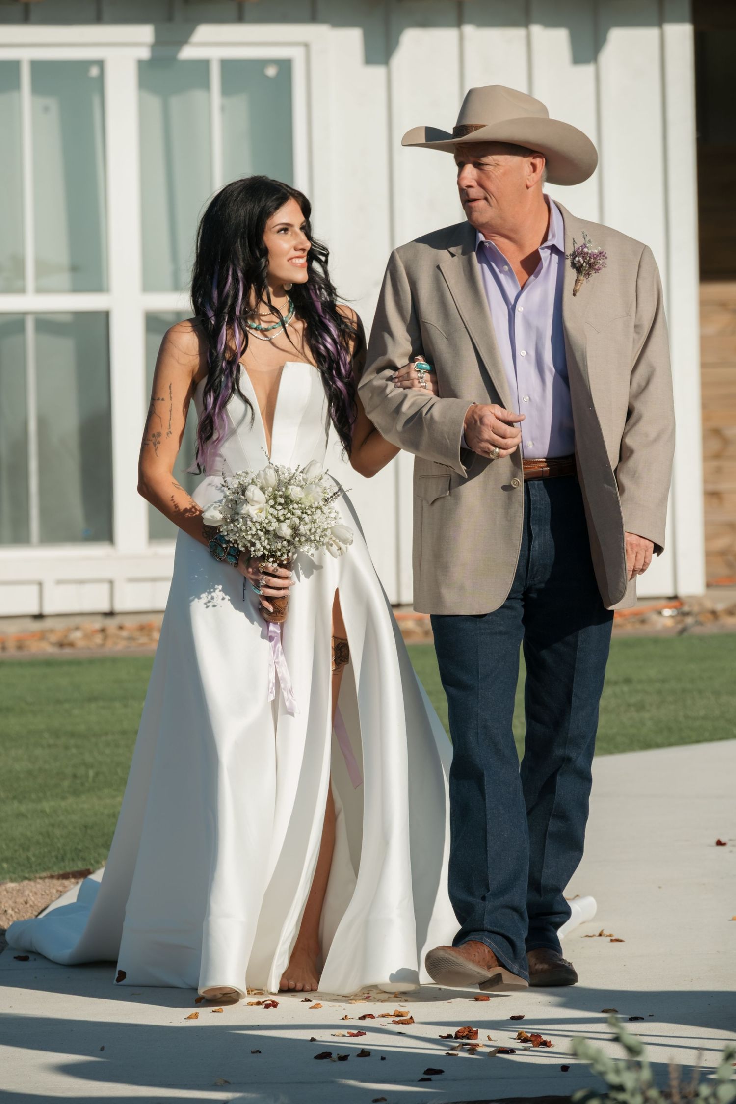 A bride in a white dress holding baby's breath bouquet walks alongside a man wearing cowboy hat and suit jacket.