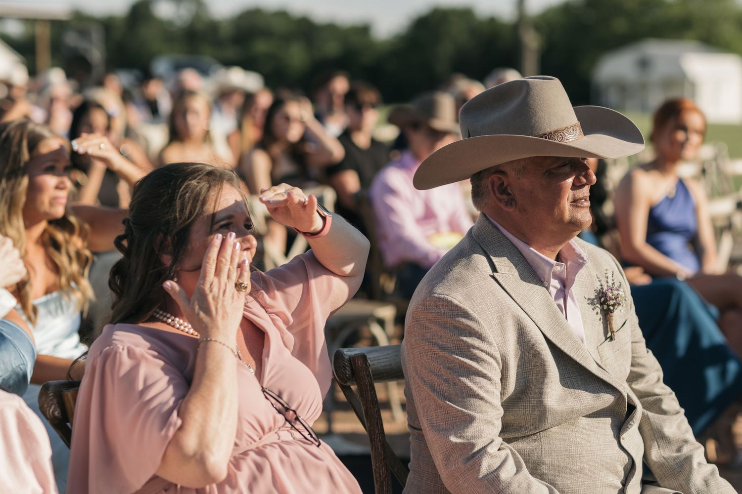 Guests sit in an outdoor audience, wearing formal attire including a cowboy hat and pink dress during a sunny day event.