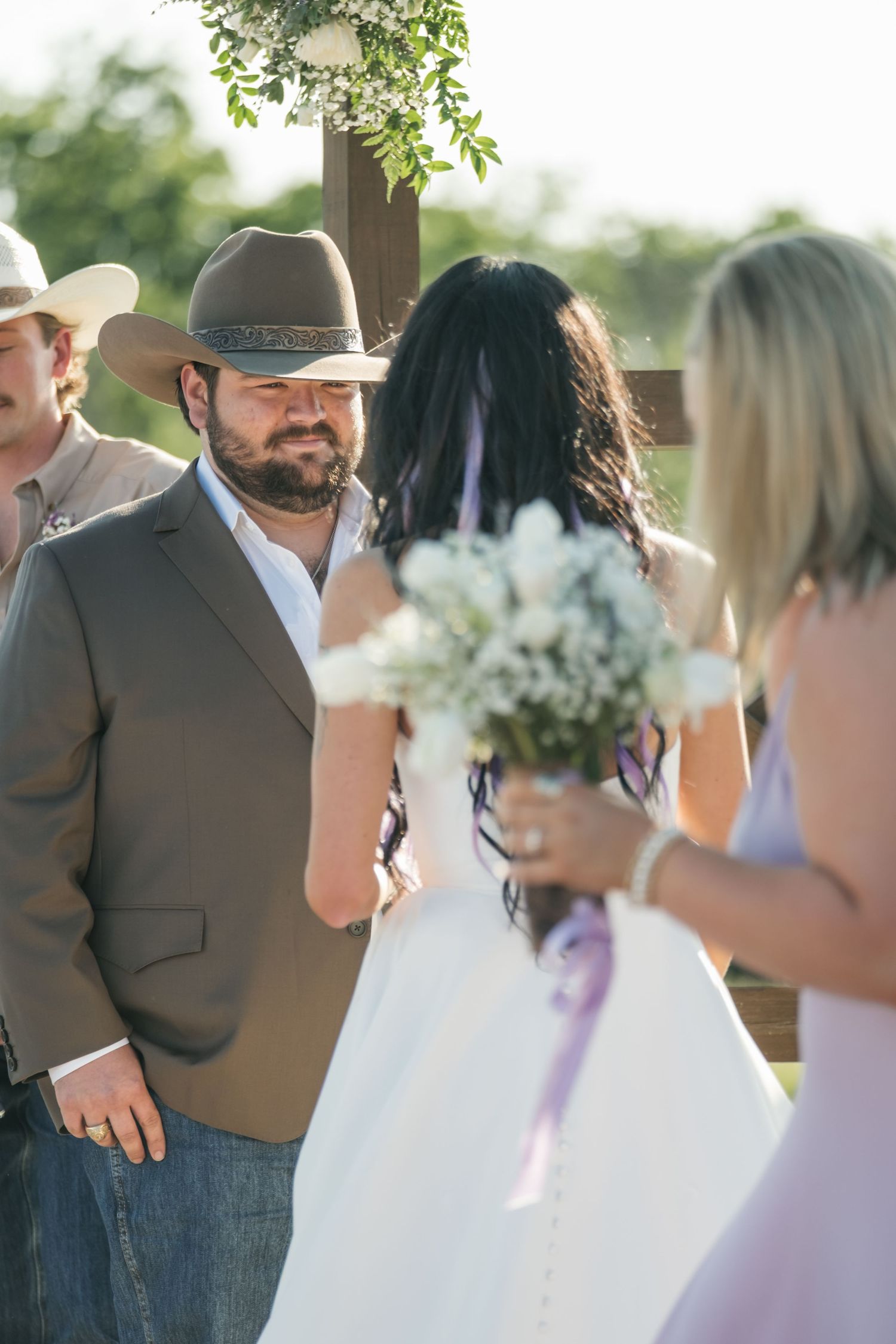 A rustic outdoor wedding ceremony features a groom in a brown jacket and white shirt standing at a wooden altar.