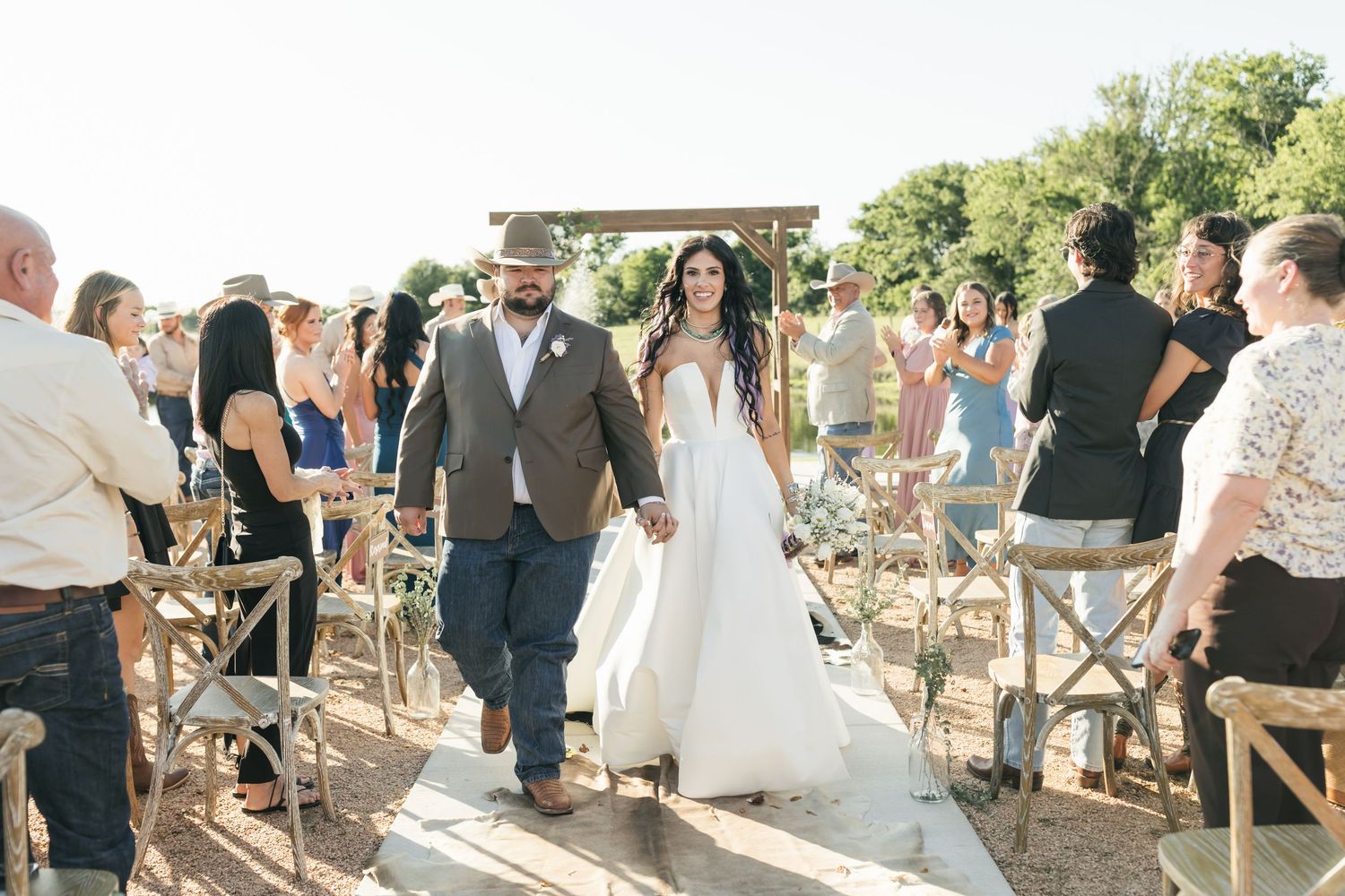 Wedding guests gather outdoors on a warm day for an informal celebration in a rustic setting with wooden chairs.