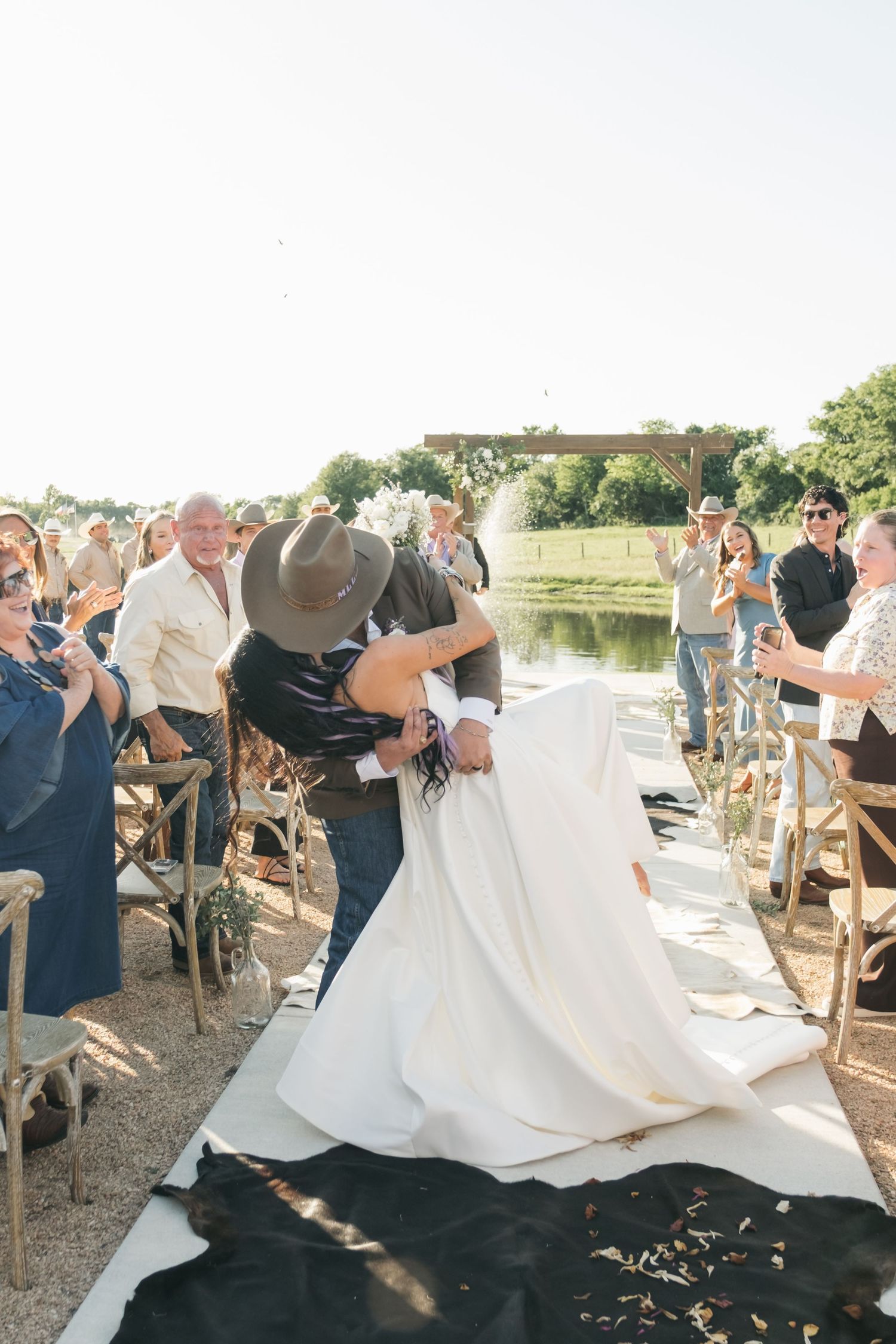 A couple shares a romantic moment during an outdoor wedding ceremony as guests look on from rustic wooden chairs.